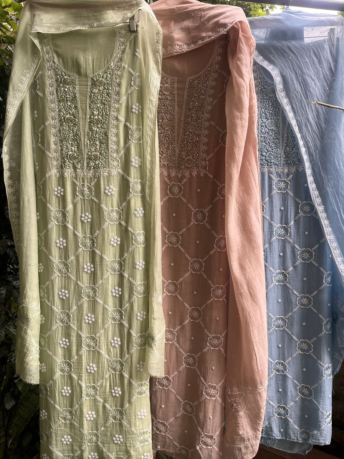 Three folded embroidered fabric garments stacked vertically, showing light blue, dusty pink, and sage green colors with white geometric and floral embroidery.