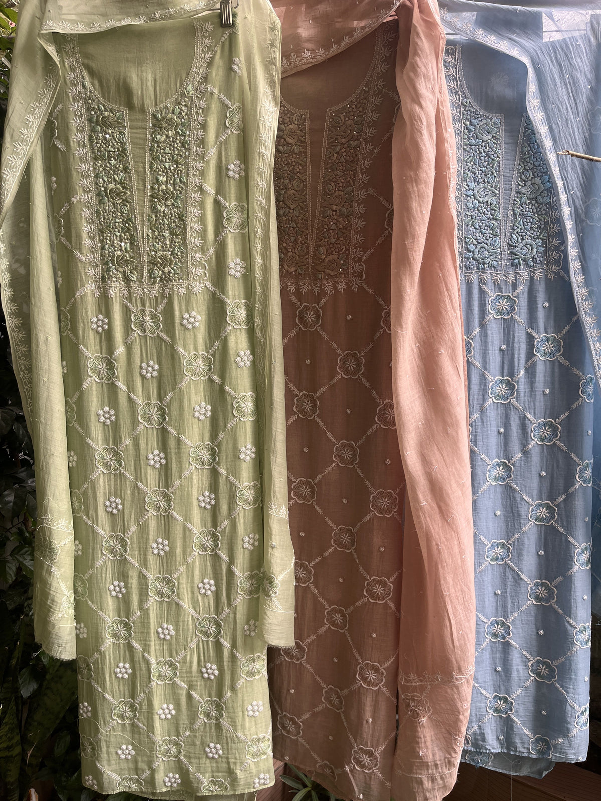 Three folded embroidered fabric pieces stacked vertically, showing different pastel shades: light blue, dusty pink, and sage green, each with white geometric and floral lace patterns.