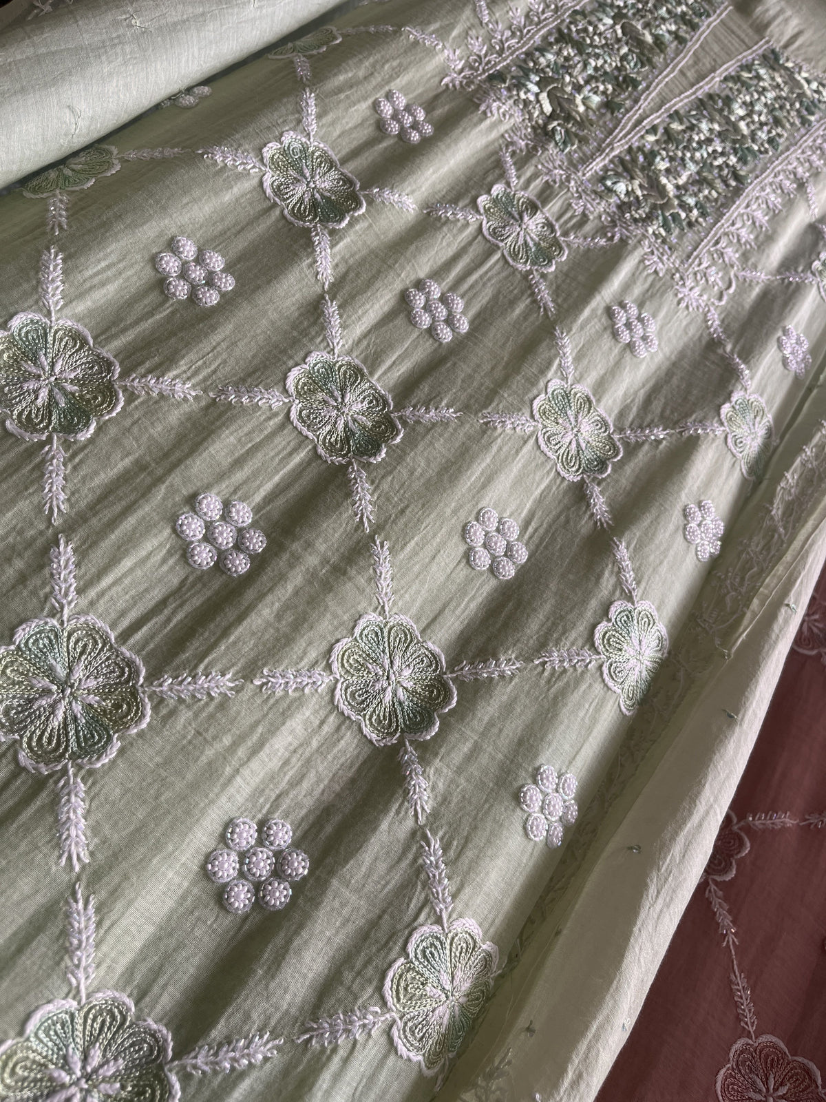 A close-up of a light sage green fabric with intricate white floral embroidery and sequin embellishments, laid out diagonally.