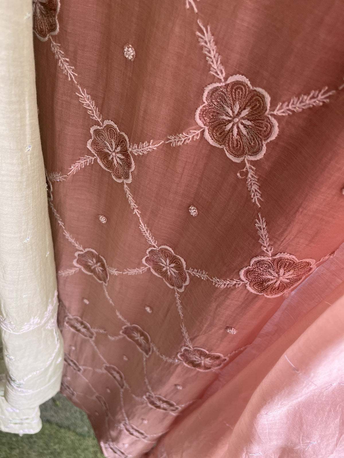 A close-up of a dusty rose pink fabric, likely a saree or dupatta, featuring intricate floral embroidery and beadwork. A light beige fabric is visible on the left edge, and a soft pink fabric is draped in the bottom right corner.