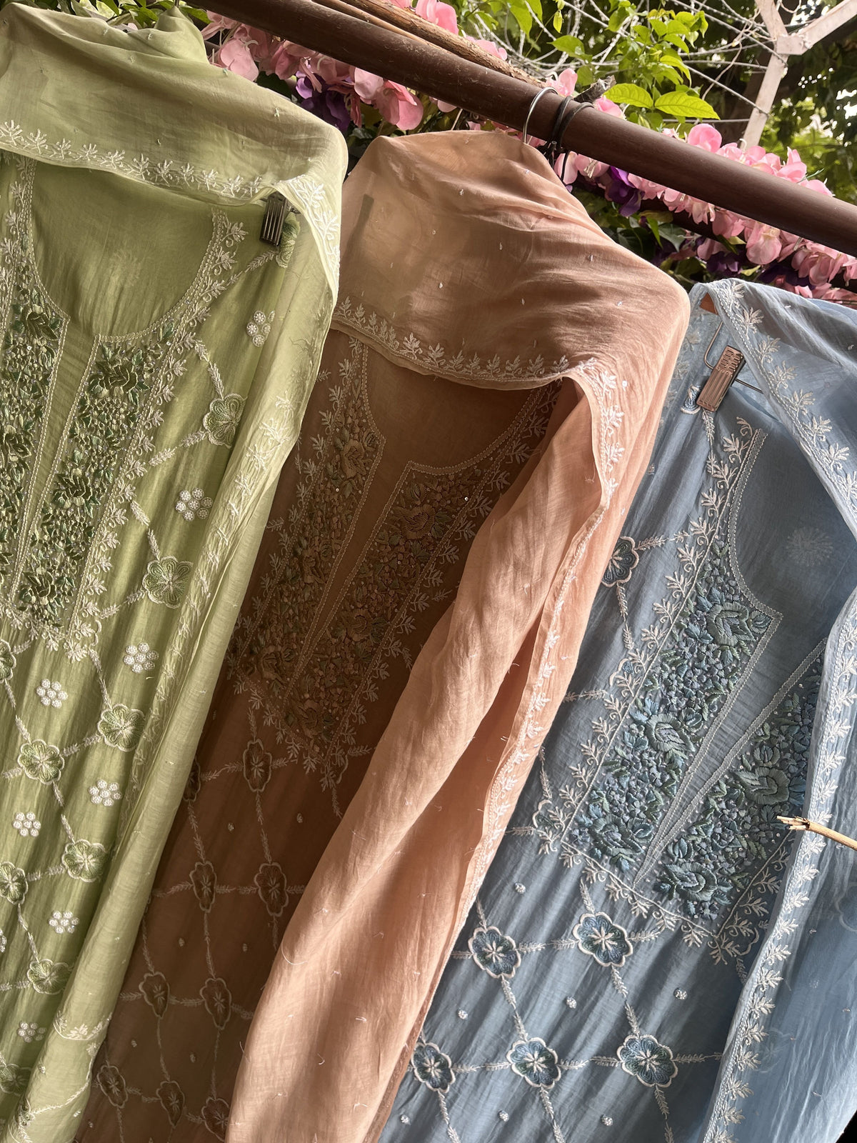 Three embroidered women's tunic dresses hanging side by side on a wooden rack outdoors. The dresses are in light green, peach, and dusty blue, each with intricate floral embroidery and beadwork on the front panels and matching dupattas.