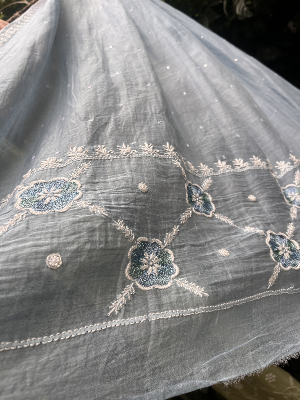 A close-up of a light gray fabric with white embroidered floral patterns and small scattered metallic embellishments.