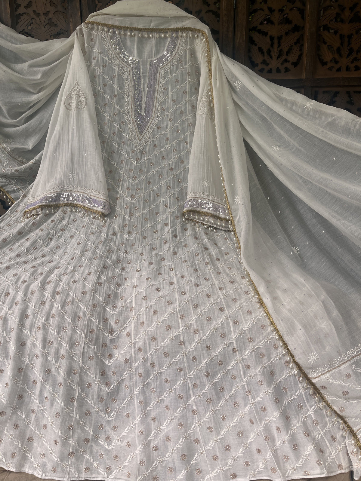 An ivory white embroidered traditional Indian outfit with a matching sheer dupatta, featuring intricate silver and gold embroidery, displayed against a dark carved wooden background.