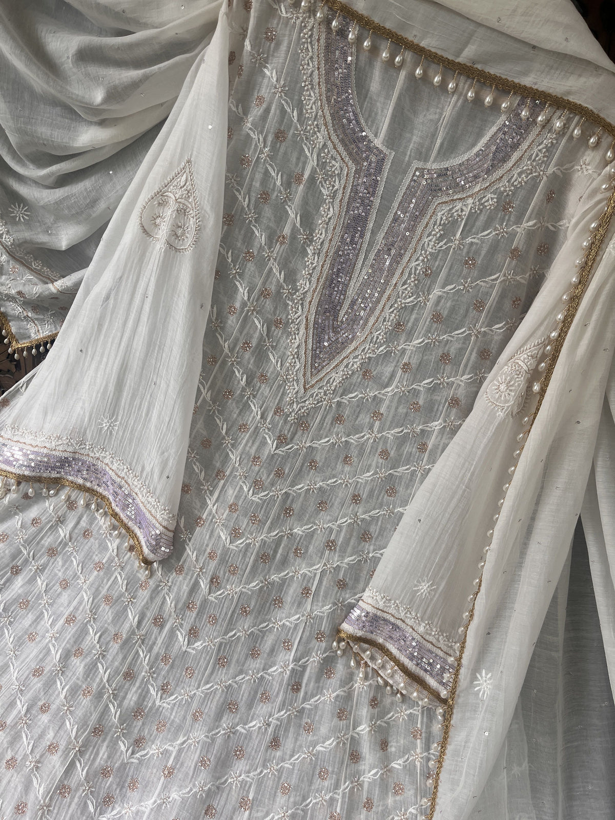 A close-up of a white or off-white embroidered fabric garment, likely a traditional outfit, featuring intricate metallic embroidery, sequins, and a matching sheer dupatta.