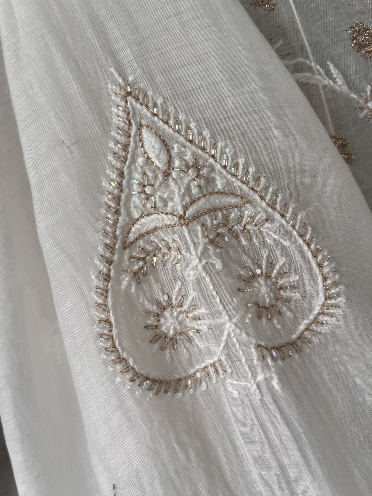 Close-up of a white or off-white embroidered fabric with a teardrop-shaped motif outlined in metallic gold thread and small floral/star details inside.