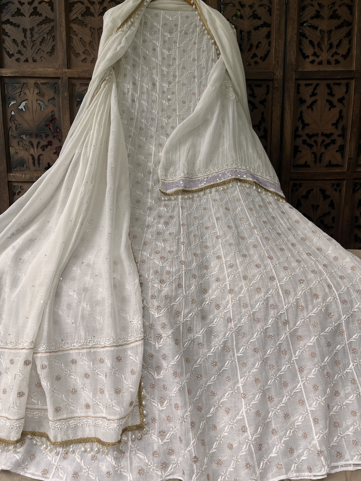 An off-white/ivory embroidered traditional outfit with a matching sheer dupatta draped over a flared skirt, set against a dark carved wooden screen.
