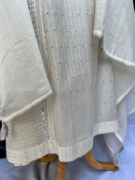 A close-up of a light beige woven textile or curtain panel with small metallic dot embellishments and a decorative silver-toned border, draped over a wooden hanger against a pale blue background.