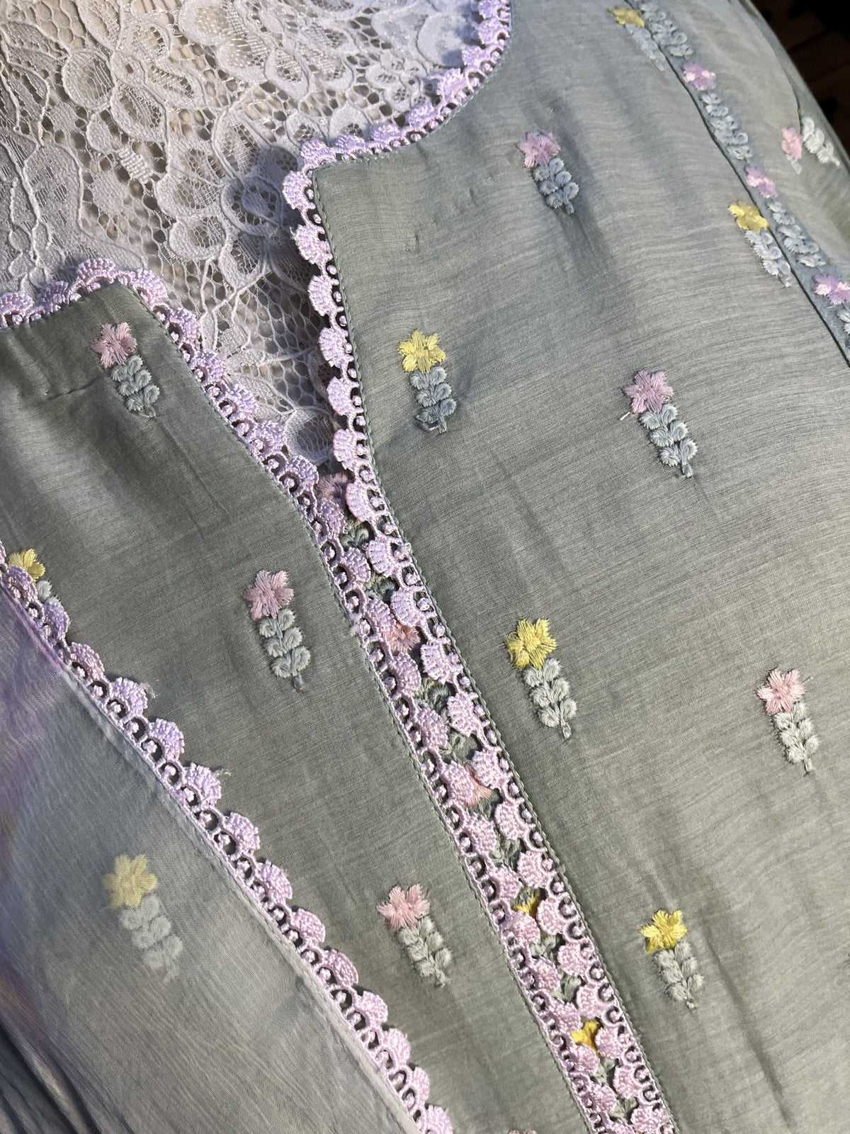 Close-up of a light gray fabric with delicate floral embroidery in pastel pink, yellow, and green, accented by a scalloped lavender lace border.