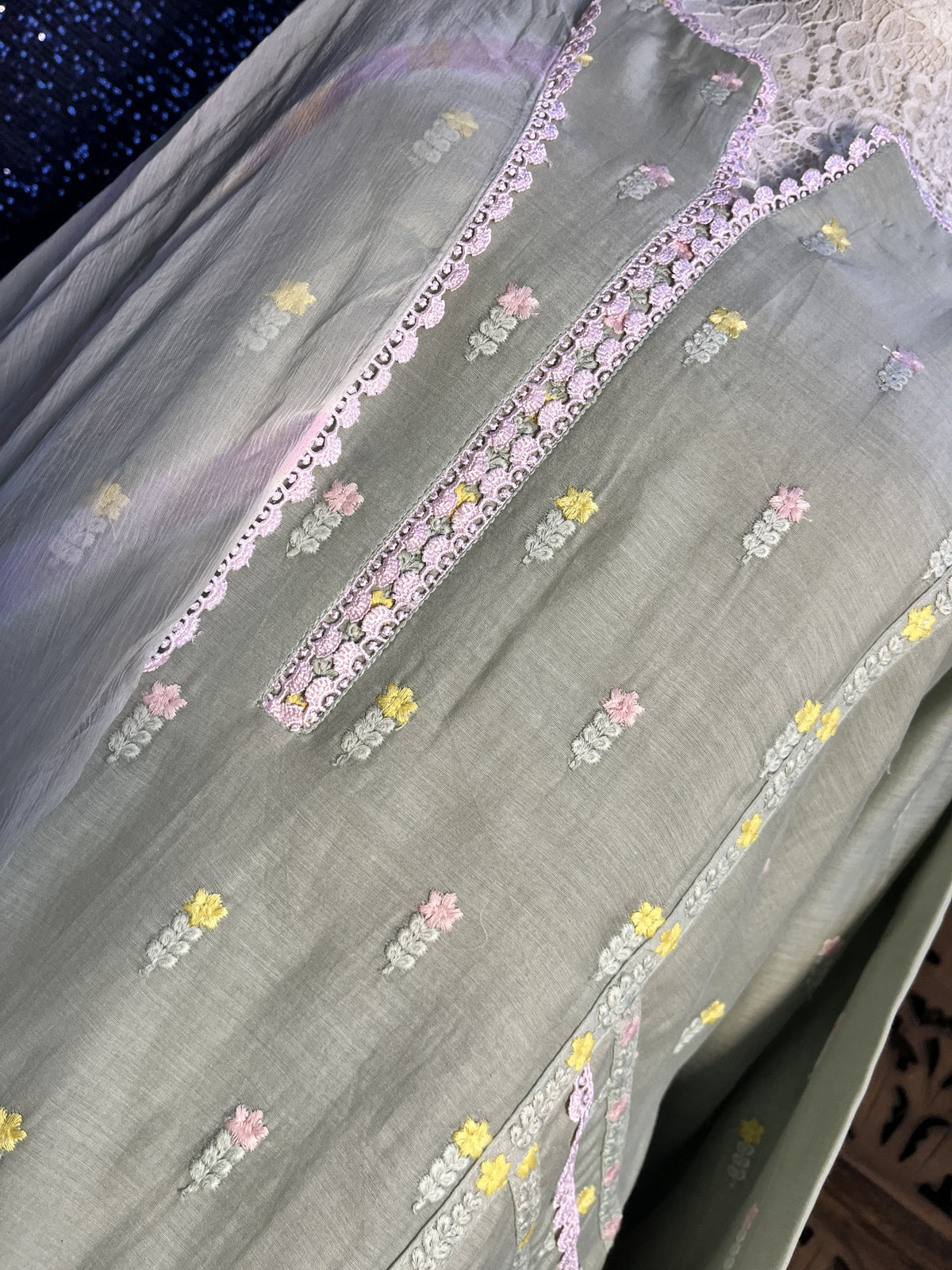 A close-up of a light sage green fabric garment or textile featuring delicate floral embroidery in pink, yellow, and white, with embroidered neckline details.