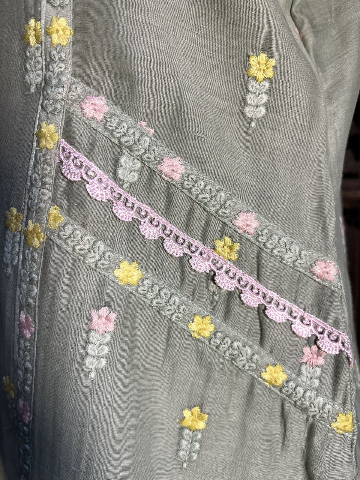 Close-up of a gray fabric garment with embroidered floral details in yellow, pink, and white, featuring decorative vertical bands of lace-like embroidery.