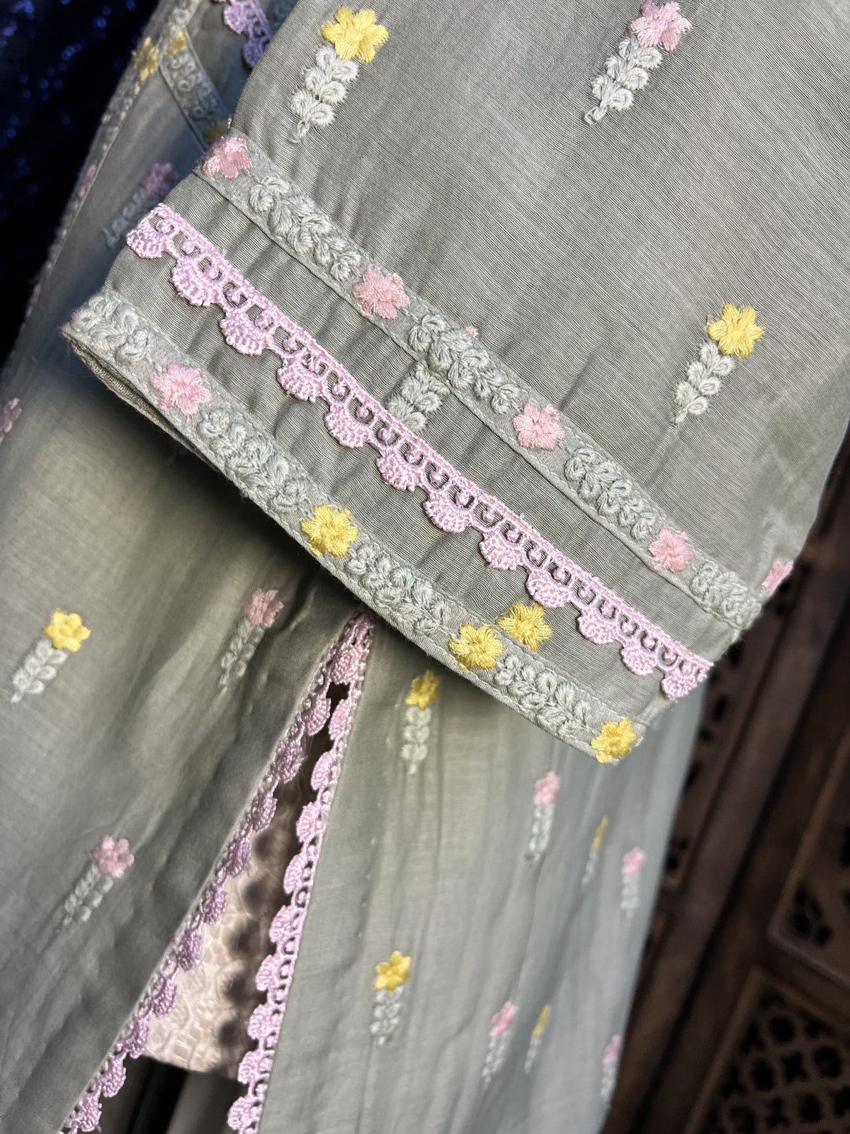 A close-up of a folded light gray fabric garment or textile with embroidered floral details in yellow, pink, and white, plus a decorative pink and gray lace border.
