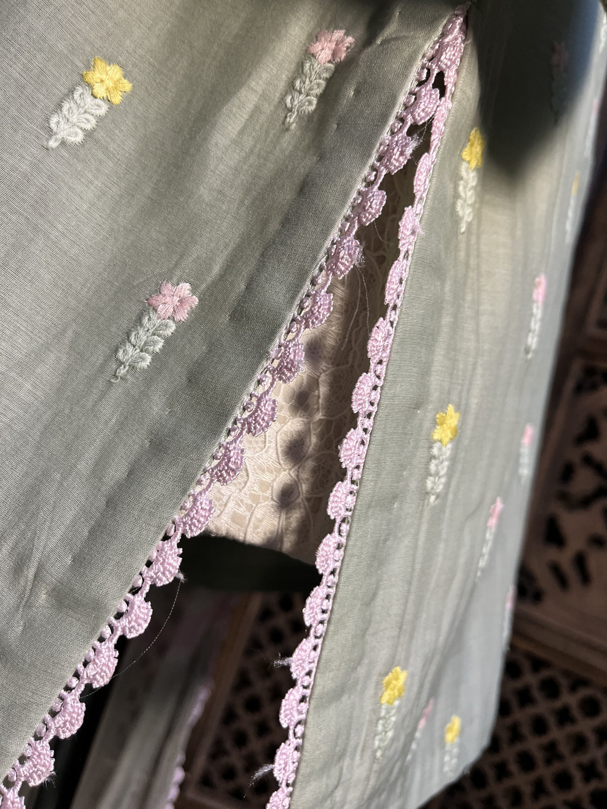 A close-up of a light sage green fabric, likely a salwar suit material, featuring embroidered floral motifs in pink, yellow, and white, finished with a pink scalloped lace border.