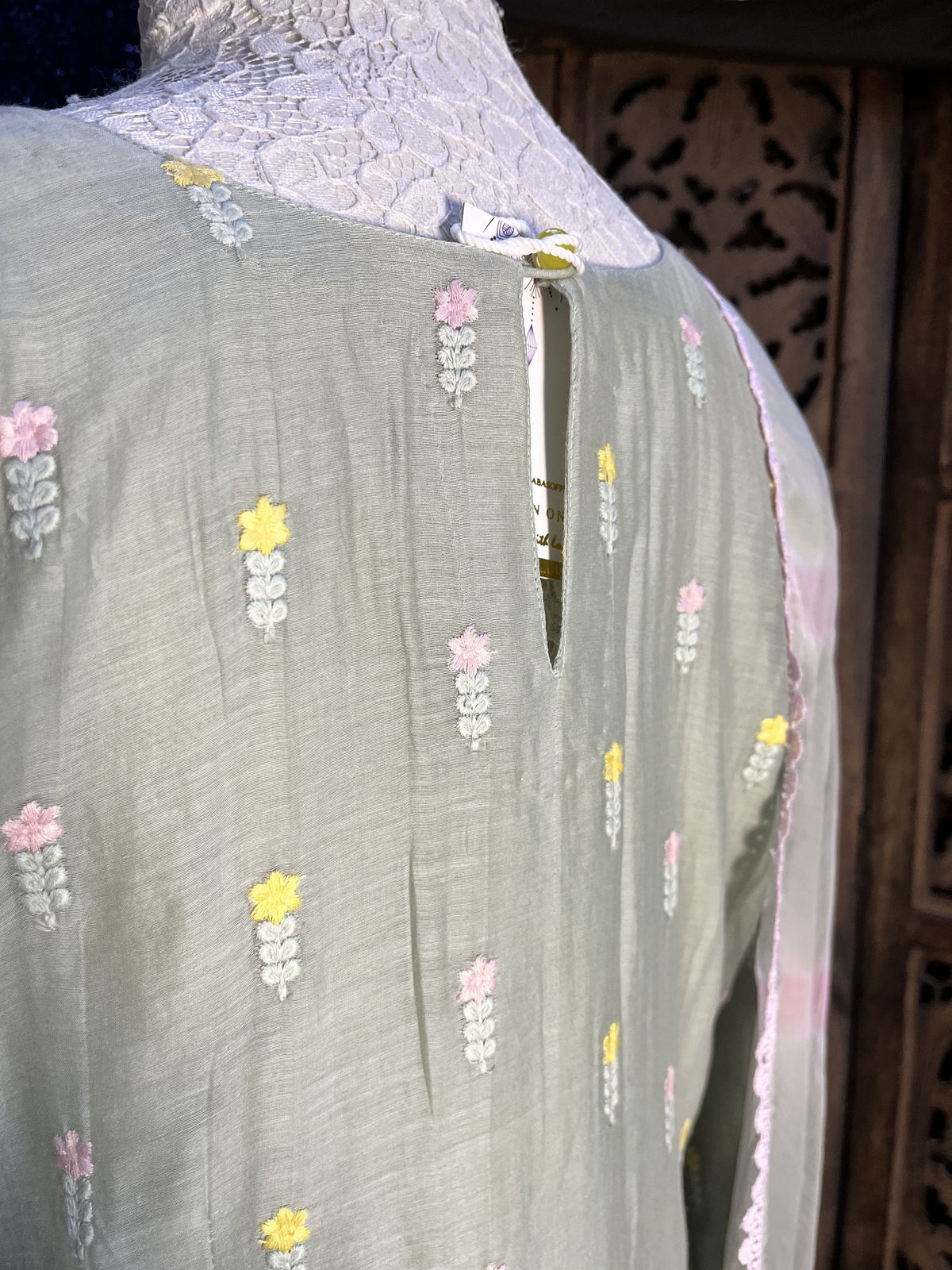 A close-up of a sage green fabric garment, likely a kurta or dress, featuring small embroidered floral motifs in pink, yellow, and white. A white lace panel is visible on the left side, and the fabric is draped over a dark wooden surface.