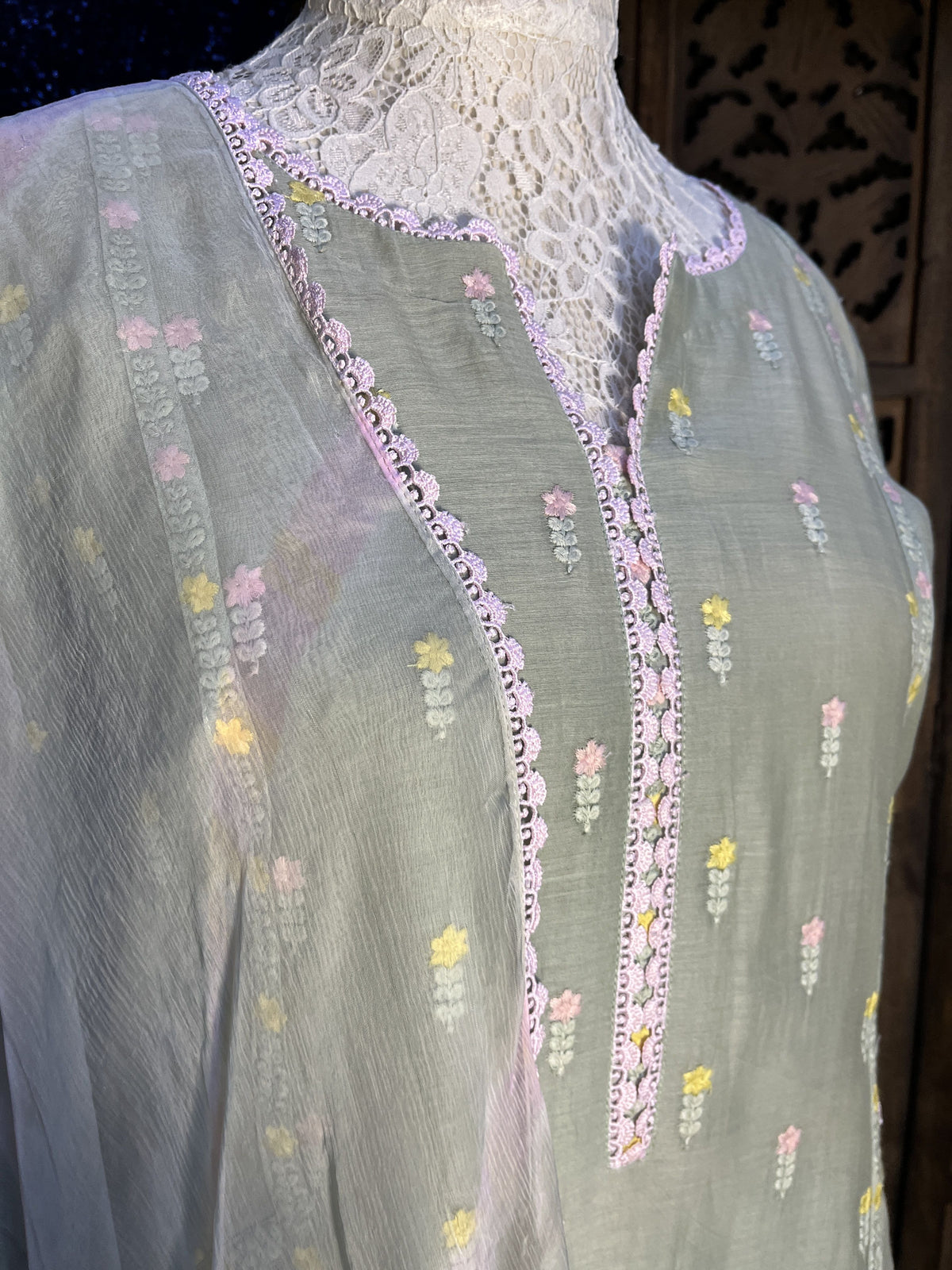 A close-up of a light sage green fabric garment or textile with embroidered floral motifs in pink, yellow, and white, featuring scalloped pink lace trim along curved neckline details.