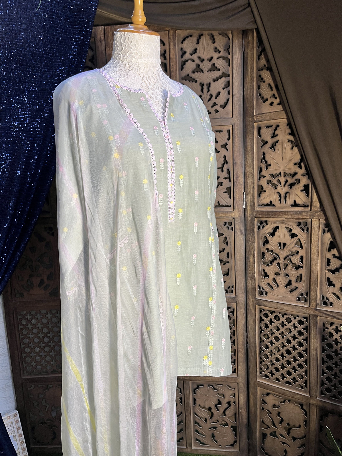 A sheer white embroidered fabric panel or scarf with small yellow and pink motifs, draped over a mannequin torso against a carved wooden background.